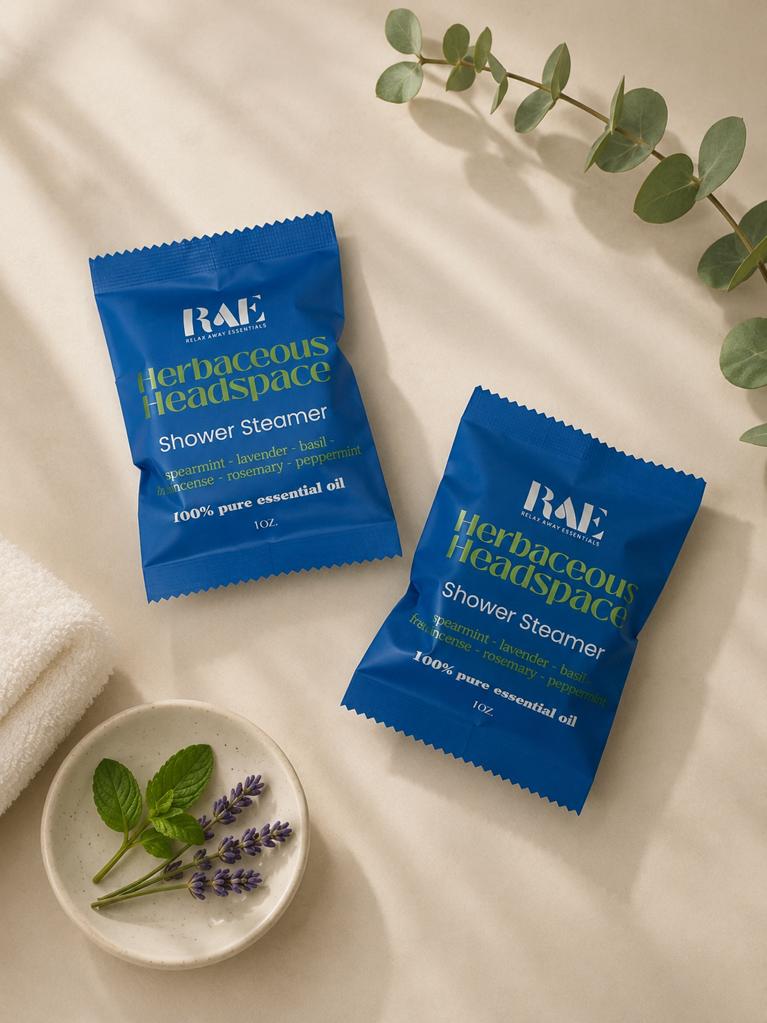 Two blue RAE Herbaceous Headspace shower steamer packets on a light surface with a small dish of lavender and mint leaves.