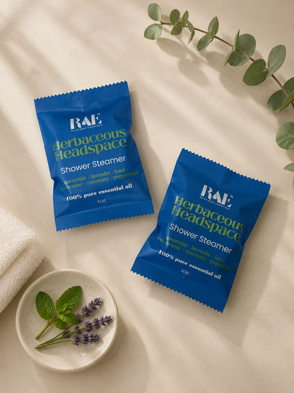 Two blue RAE Herbaceous Headspace shower steamer packets on a light surface with a small dish of lavender and mint leaves.