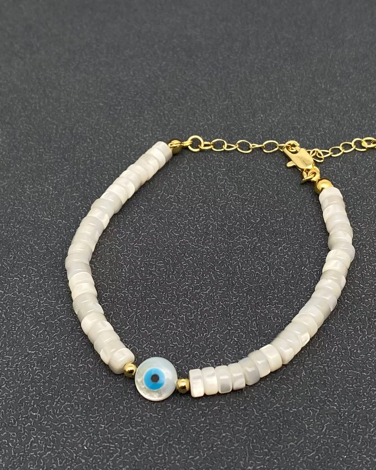 A beaded bracelet featuring white disc-shaped beads, a central blue and white evil eye charm, and gold-tone metal chain and clasp, displayed on a dark textured background.