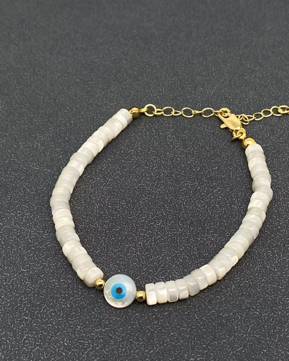 A beaded bracelet featuring white disc-shaped beads, a central blue and white evil eye charm, and gold-tone metal chain and clasp, displayed on a dark textured background.