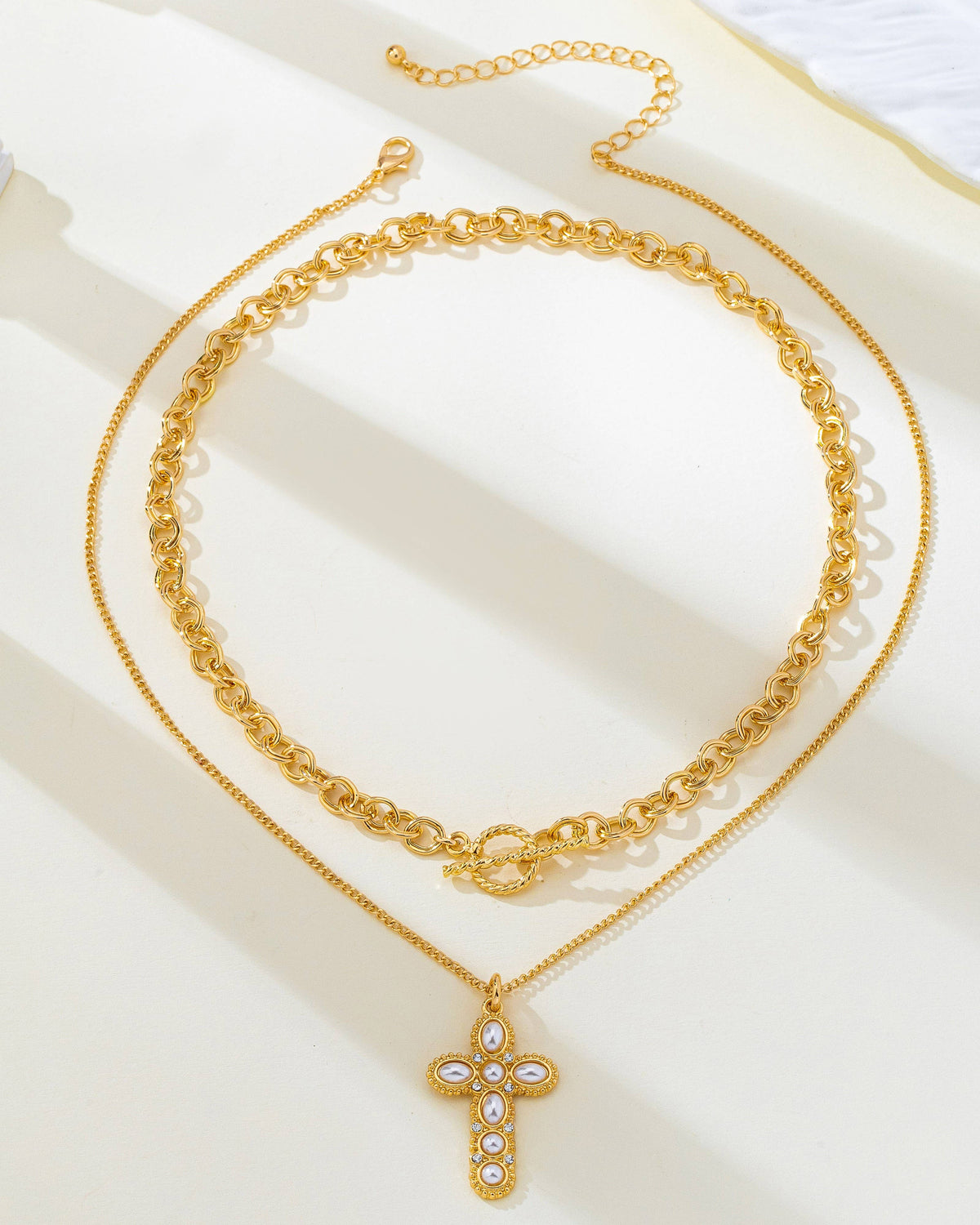A double-layered gold necklace set featuring a thicker curb chain and a slimmer cable chain with a cross pendant adorned with pearls and rhinestones, displayed on a cream-colored surface with soft shadows.