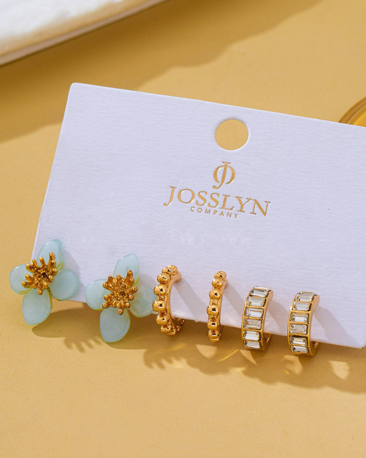 A set of three gold-toned earrings displayed on a white jewelry card, including two flower-shaped studs with pale green resin petals and two gold hoop styles, photographed on a warm beige surface.