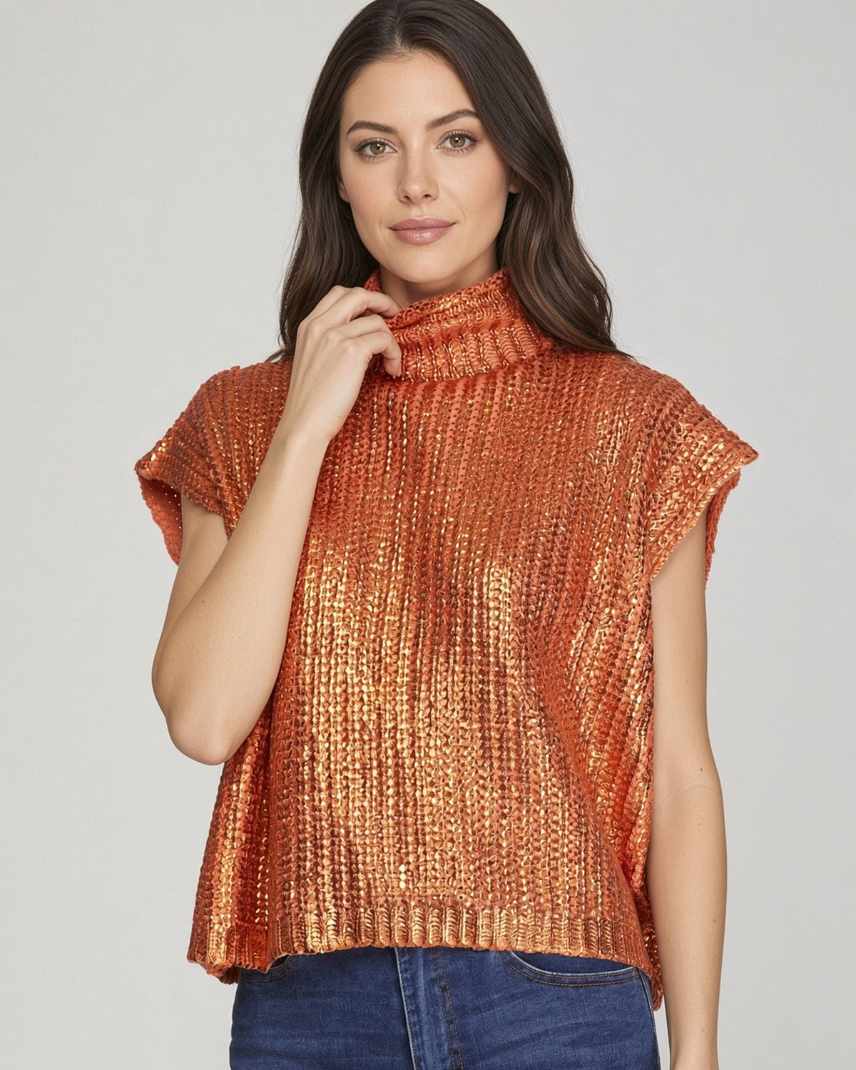 Model wearing a blaze orange heavy knit metallic sweater top with mock neck and short sleeves with blue jeans.