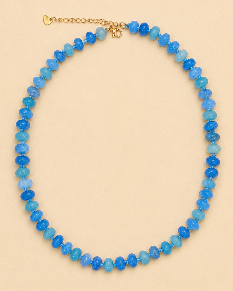 A beaded necklace made of faceted blue and light blue beads with a gold-tone clasp, displayed on a solid beige background.