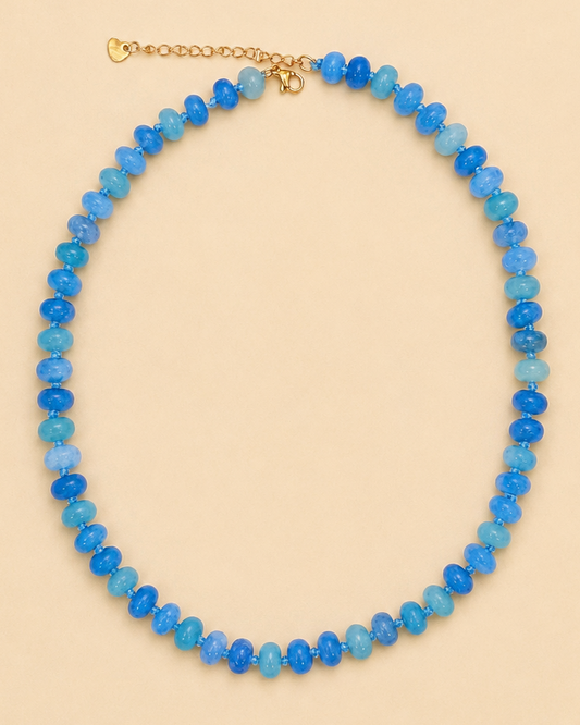 A beaded necklace made of faceted blue and light blue beads with a gold-tone clasp, displayed on a solid beige background.
