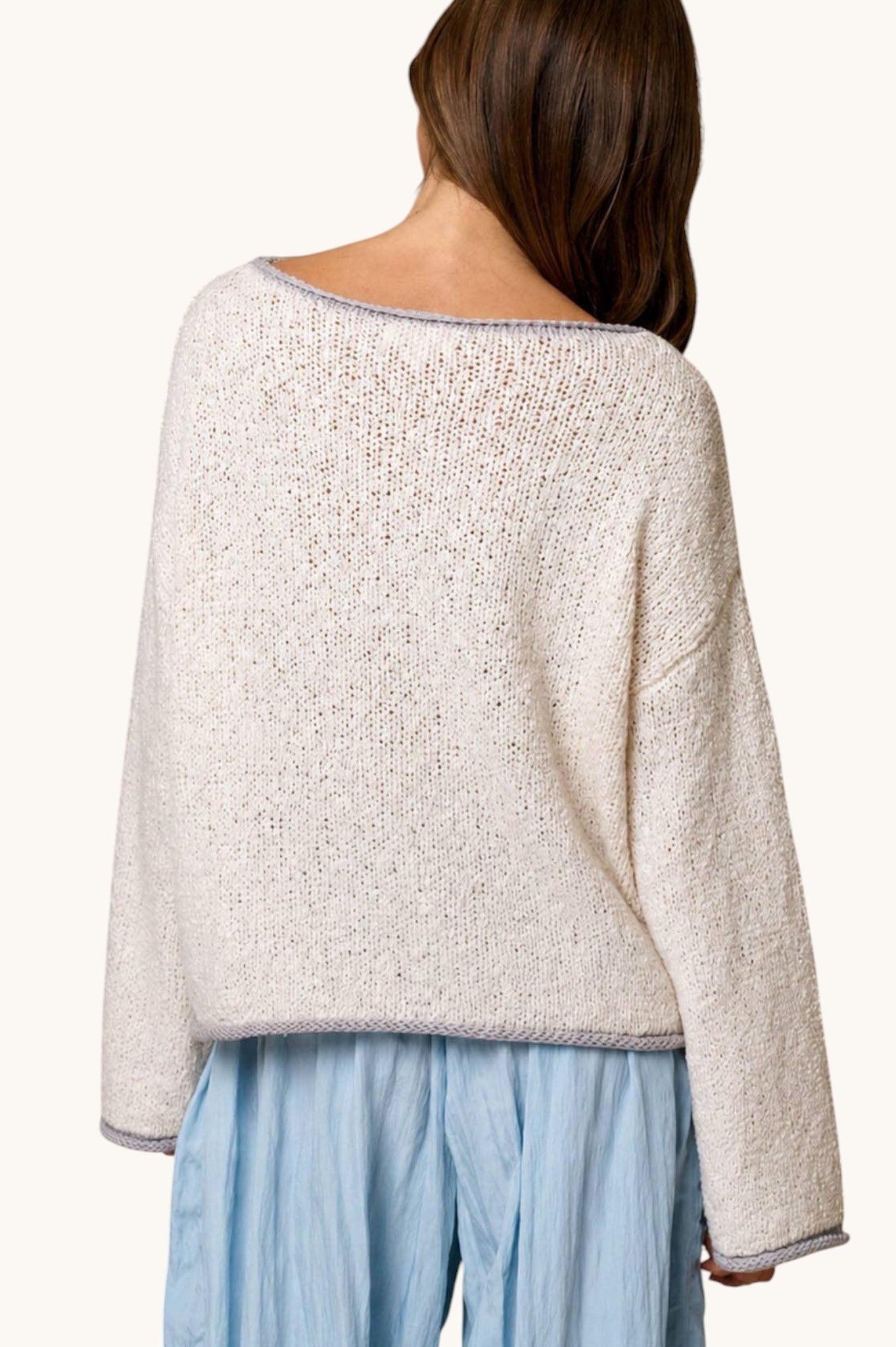 A woman shown from the back wearing a loose, textured knit sweater in a light cream or off-white color, paired with light blue pants, against a plain light background.