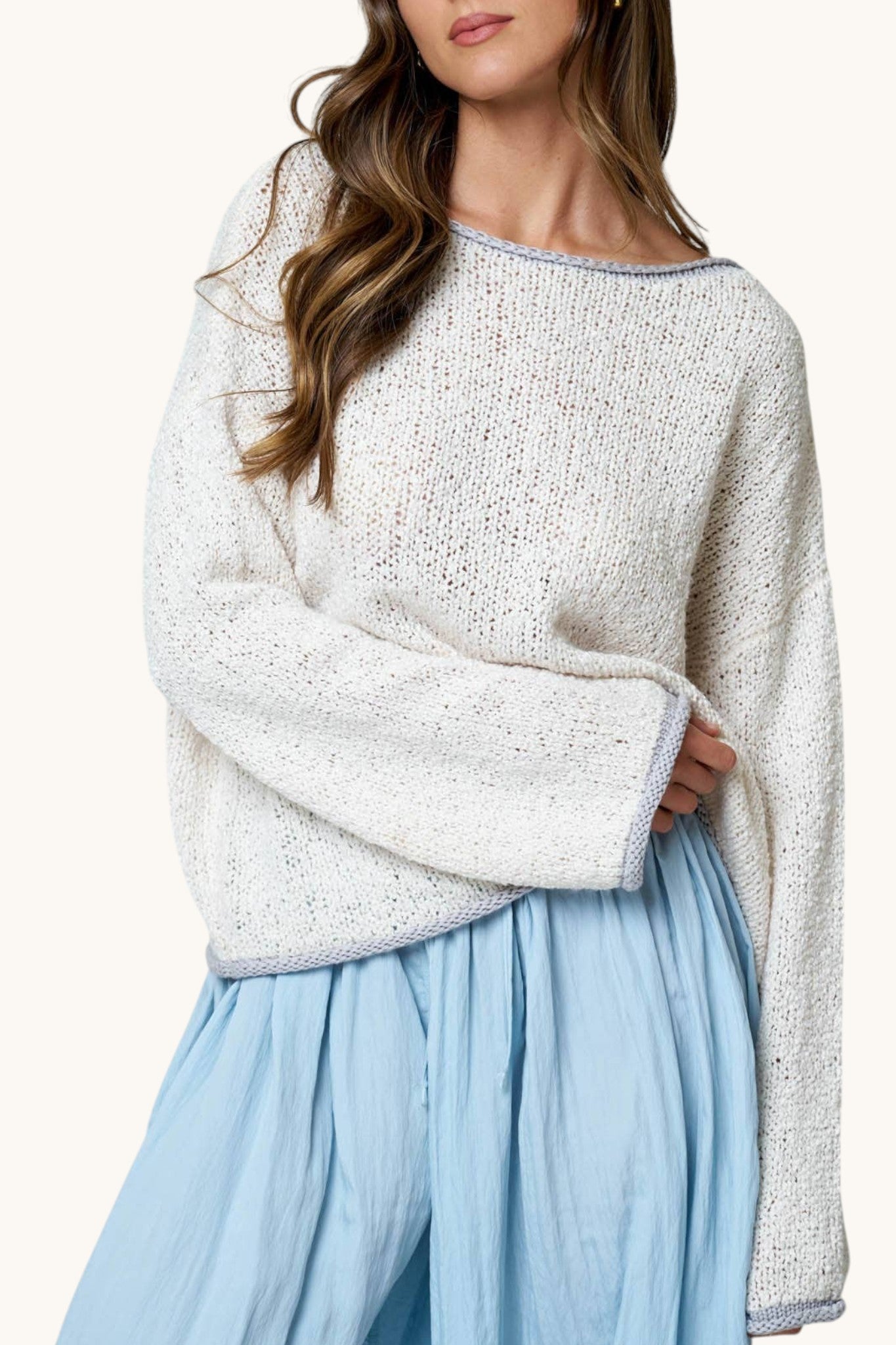 A woman wearing a chunky knit off-white sweater with a light grey trim and a light blue skirt, shown from the neck down against a plain light background.