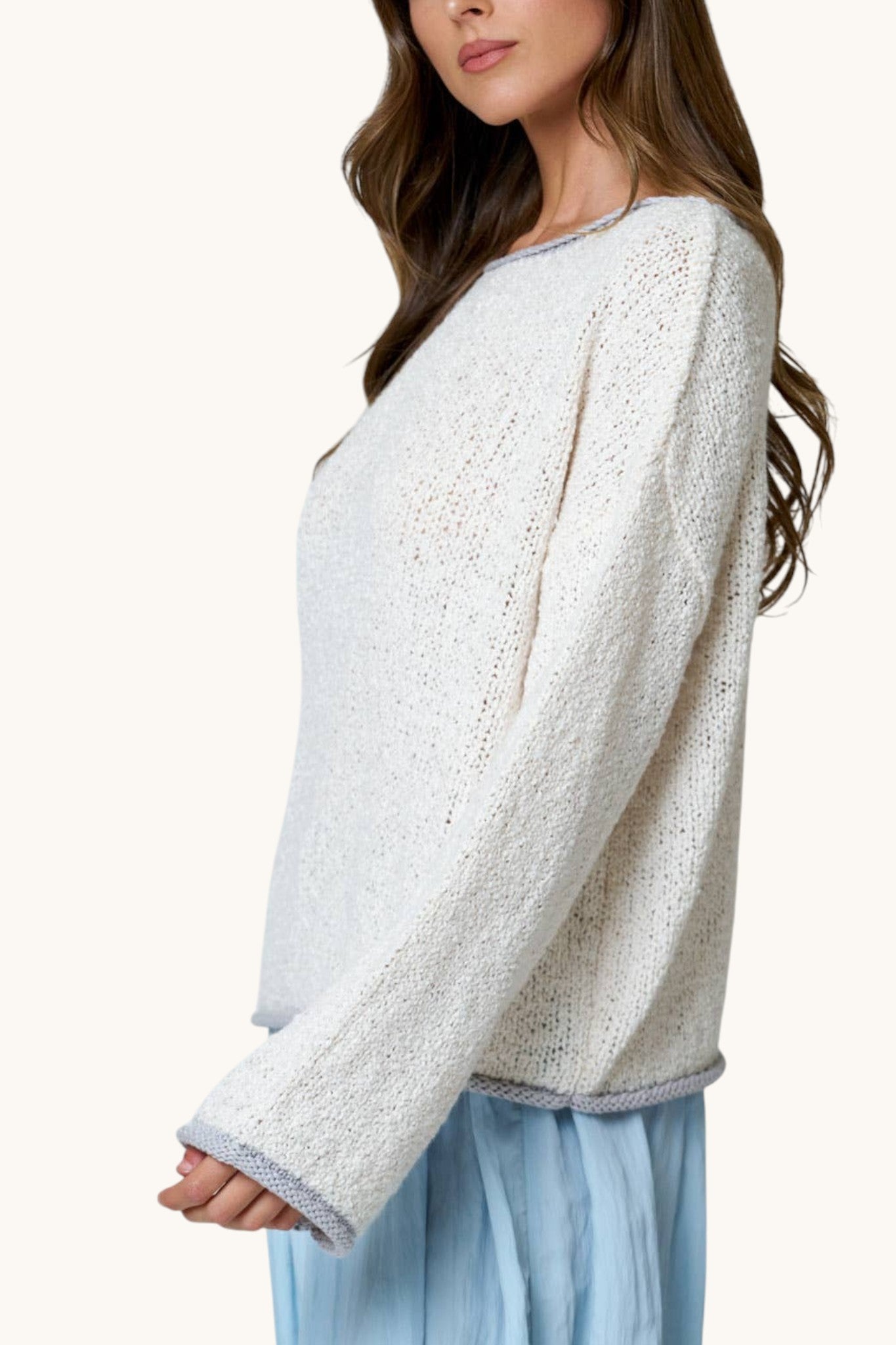 A woman wearing a cream-colored, textured open-knit sweater with light gray trim, paired with light blue bottoms, shown from a side profile against a solid off-white background.