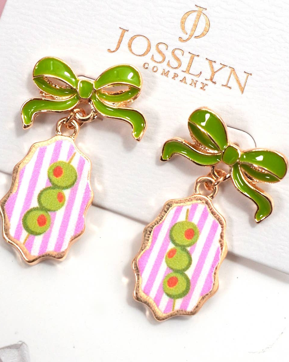 A pair of Josslyn Company enamel drop earrings displayed on a white branded card. Each earring has a glossy green bow at the top and a dangling shield-shaped charm with pink and white vertical stripes, three green olives and outlined in gold-tone metal.