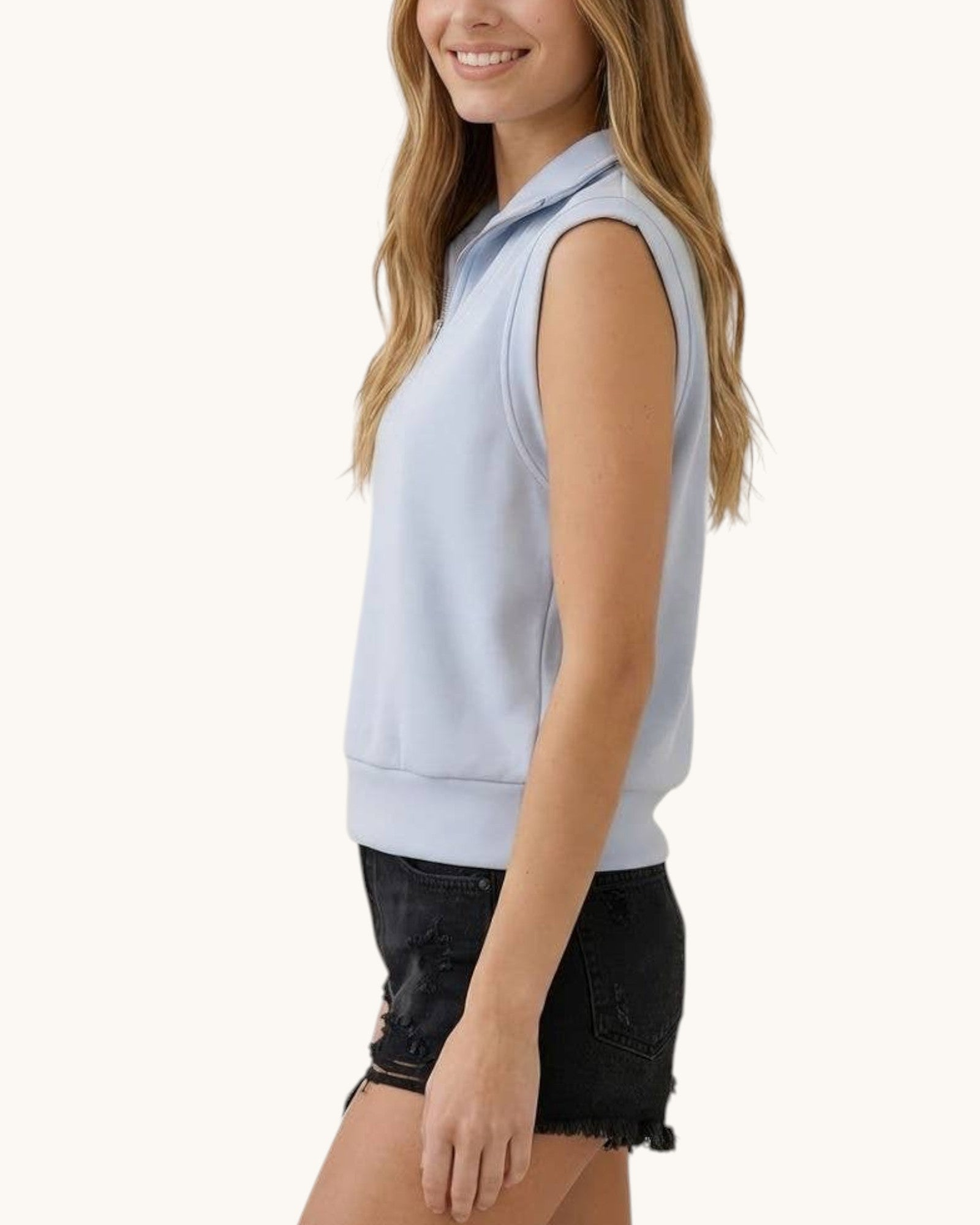 A woman wearing a light blue sleeveless sweatshirt vest and black denim shorts, shown from a side profile against a plain off-white background.