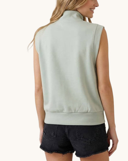 A woman shown from the back wearing a sleeveless high-neck athleisure top in a pale sage green color, paired with dark denim shorts, against a plain off-white background.