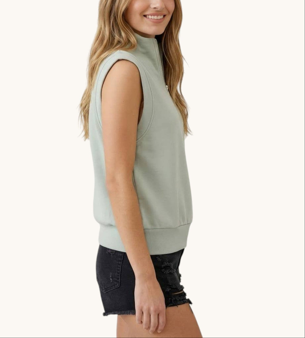 A woman wearing a sage green sleeveless mock neck modal sweatshirt and dark denim shorts, shown from a side profile against a solid off-white background.