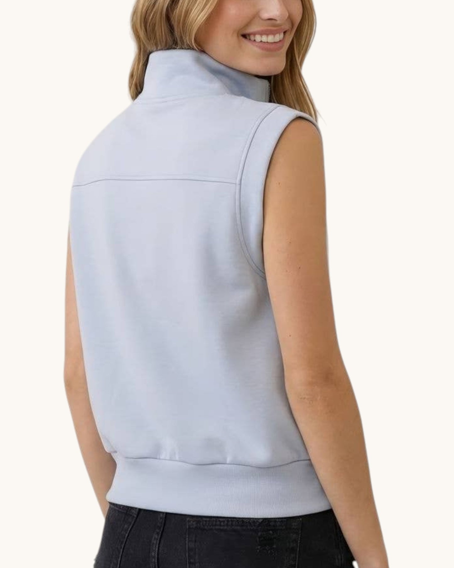 A woman shown from the back wearing a sleeveless light blue mock neck vest/top, paired with dark jeans, against a plain off-white background.