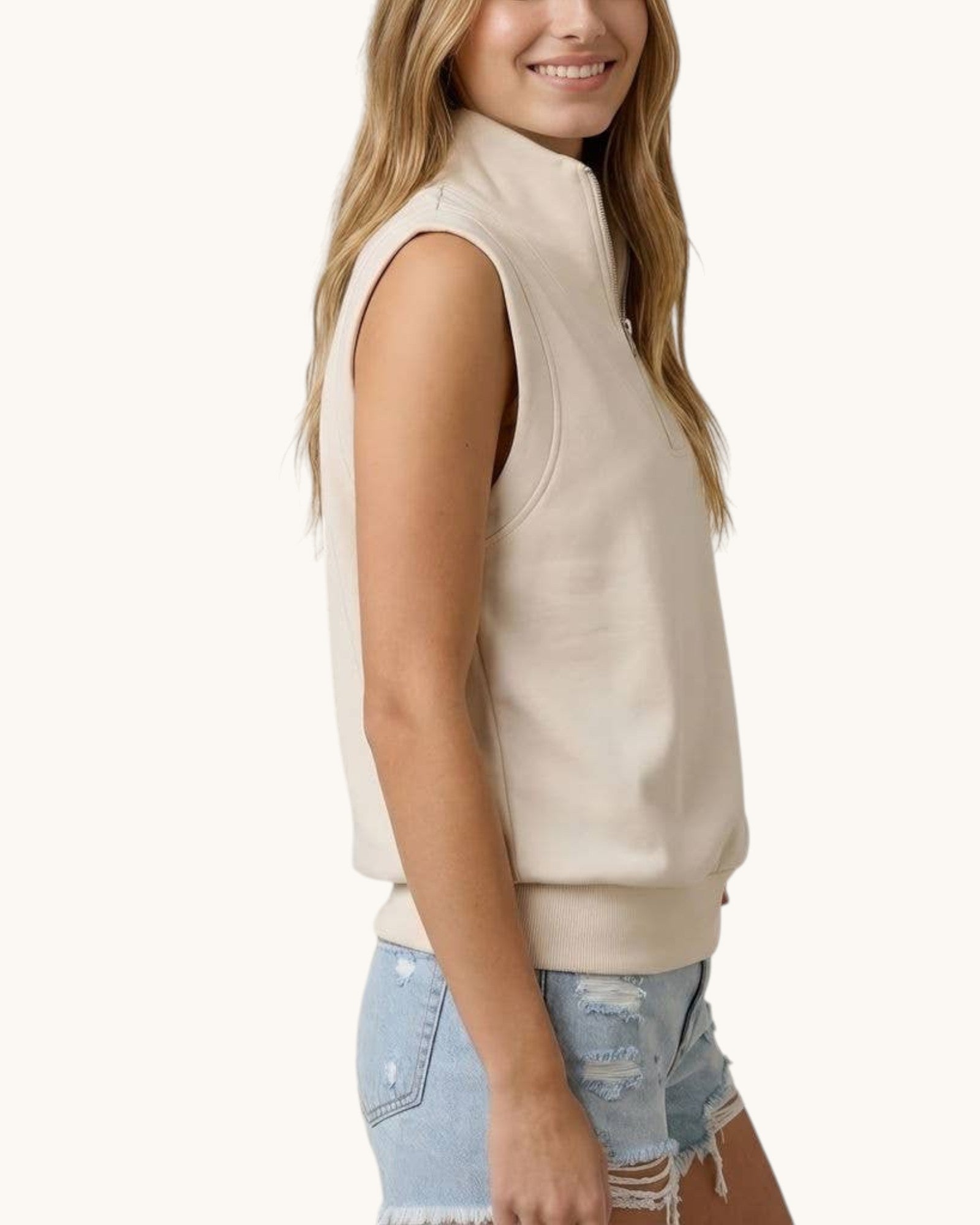 Side view of a woman wearing a ivory sleeveless jacket and denim shorts on a white background.