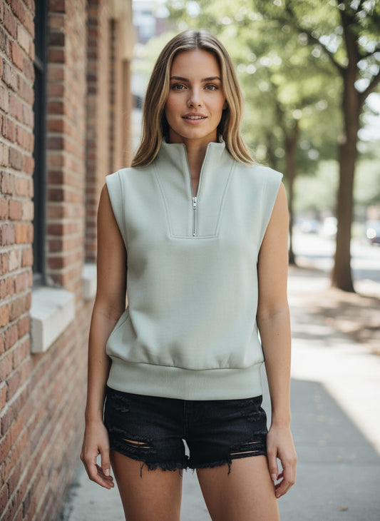 A woman wearing a sage green sleeveless quarter-zip pullover top and black distressed denim shorts, standing outdoors on a sidewalk next to a brick wall.