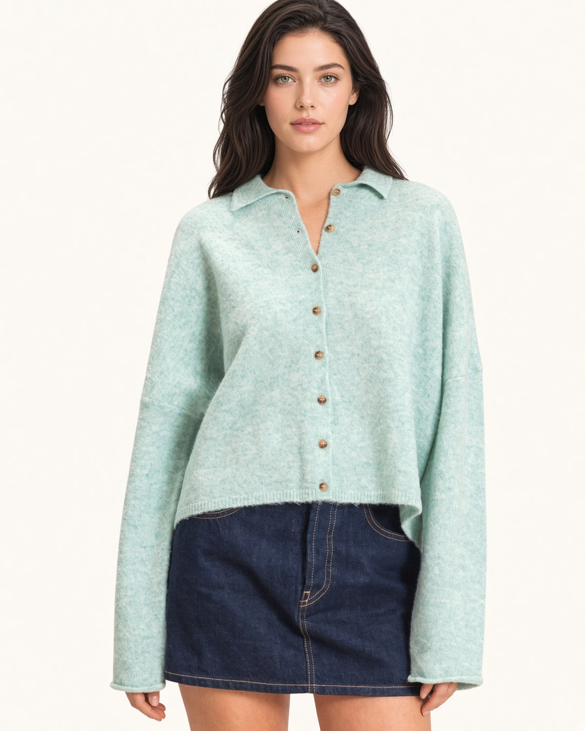 A woman modeling a light mint green knit button-up cardigan with brown buttons, paired with a dark denim mini skirt, against a plain off-white background.