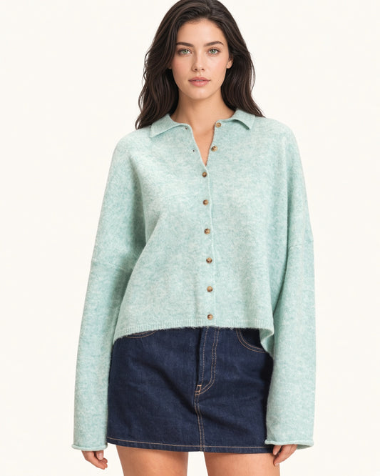 A woman modeling a light mint green knit button-up cardigan with brown buttons, paired with a dark denim mini skirt, against a plain off-white background.