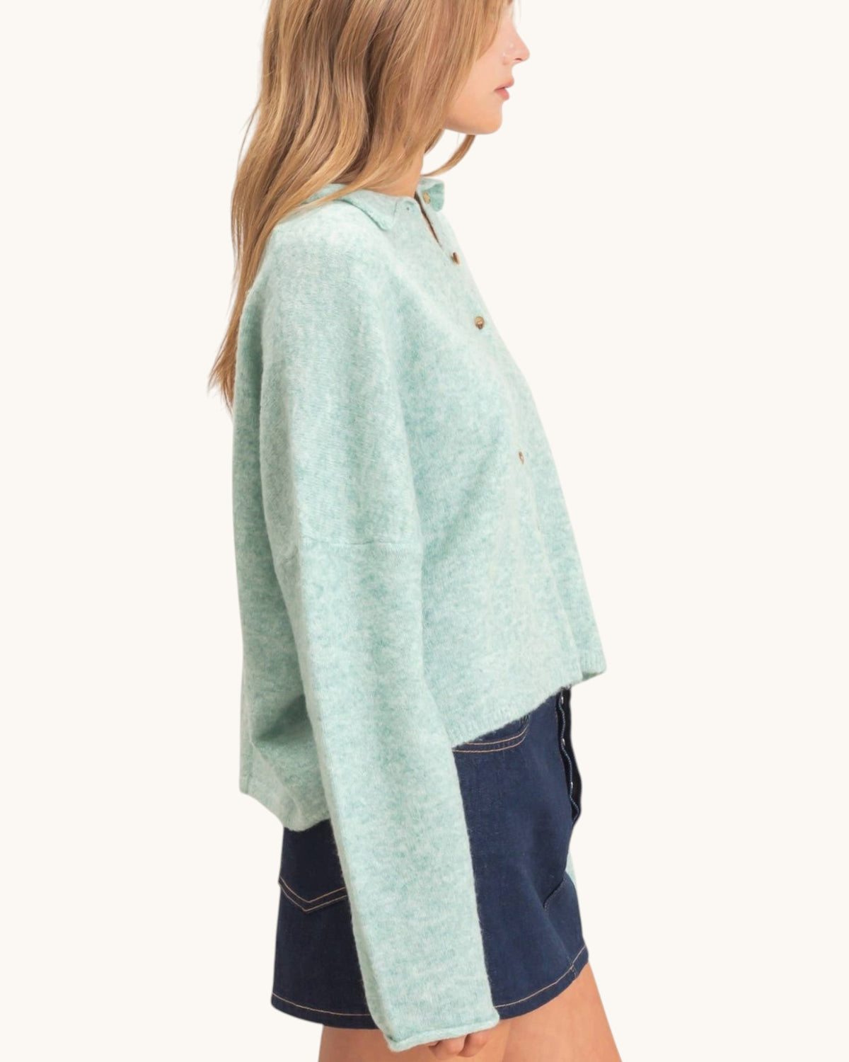 A woman shown in side profile wearing a light mint green cropped button-front cardigan or sweater with long sleeves, paired with dark denim shorts, against a plain off-white studio background.