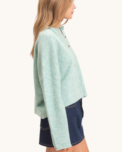 A woman shown in side profile wearing a light mint green cropped button-front cardigan or sweater with long sleeves, paired with dark denim shorts, against a plain off-white studio background.