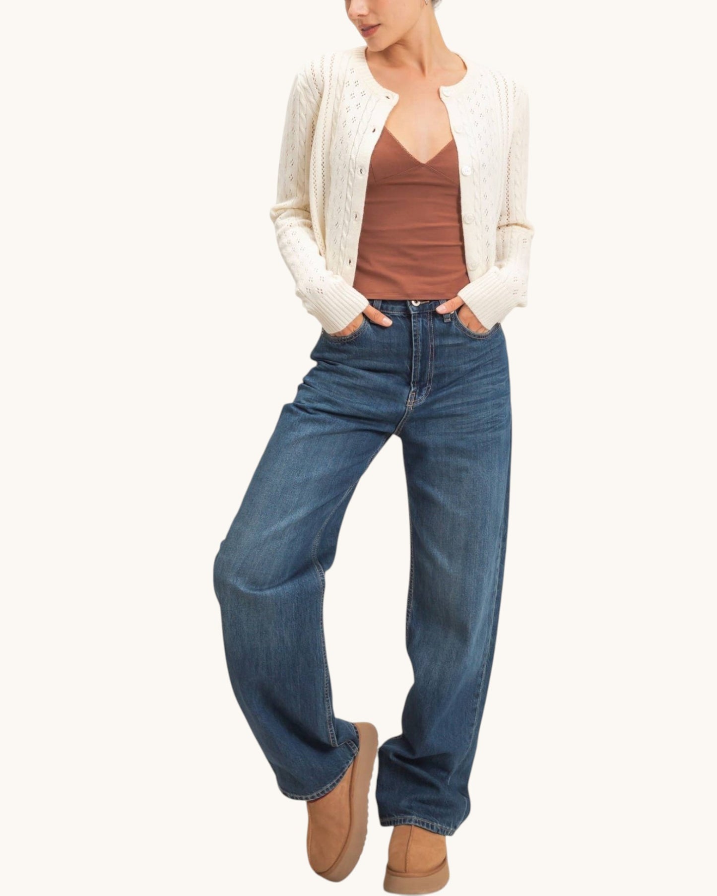 A model wearing a cream cable-knit cardigan over a brown camisole, paired with wide-leg blue jeans and tan boots, against a solid off-white background.