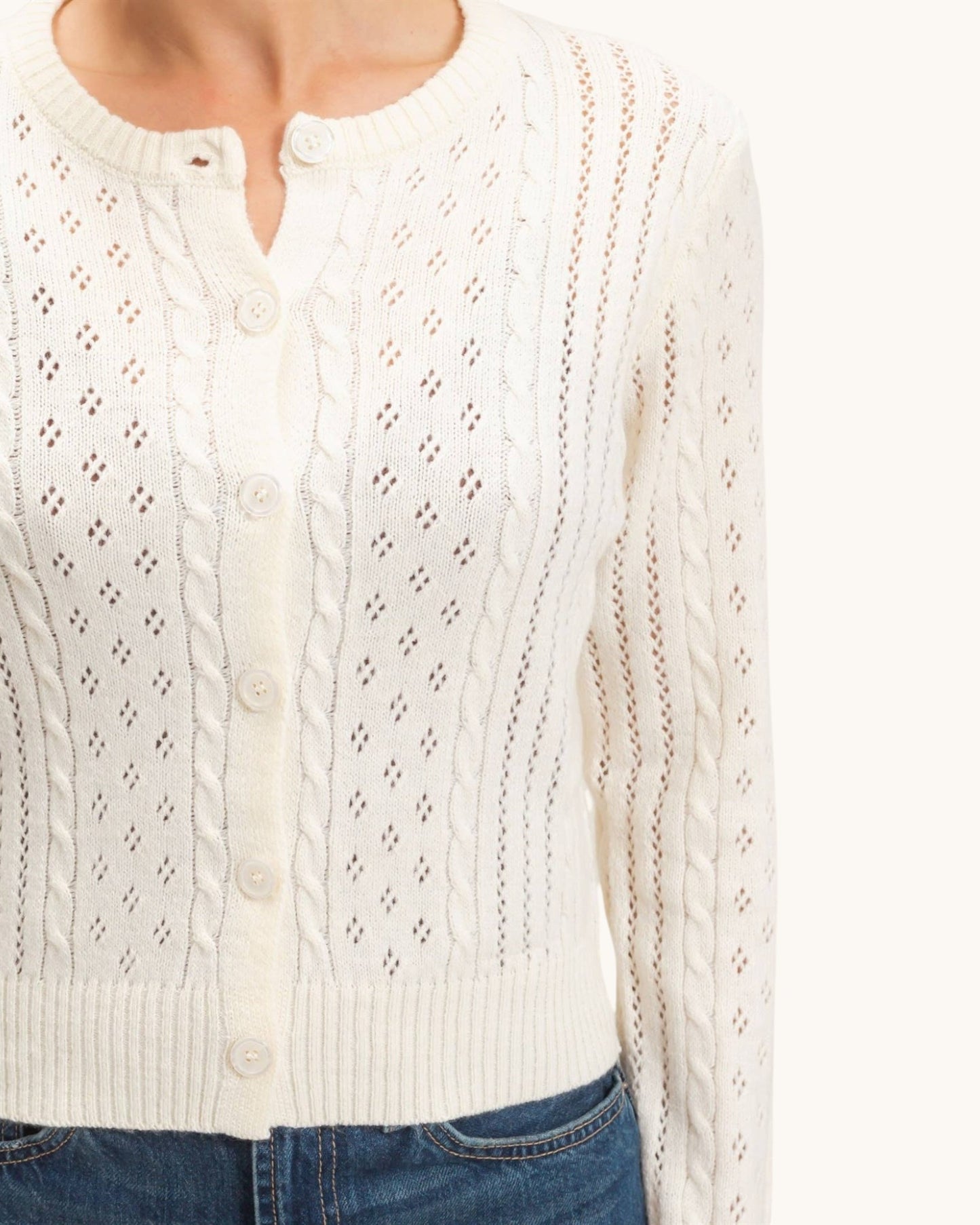 An close up of an off-white cable-knit cardigan sweater with button-front closure and openwork knit details, shown on a model with blue jeans against a plain light background.