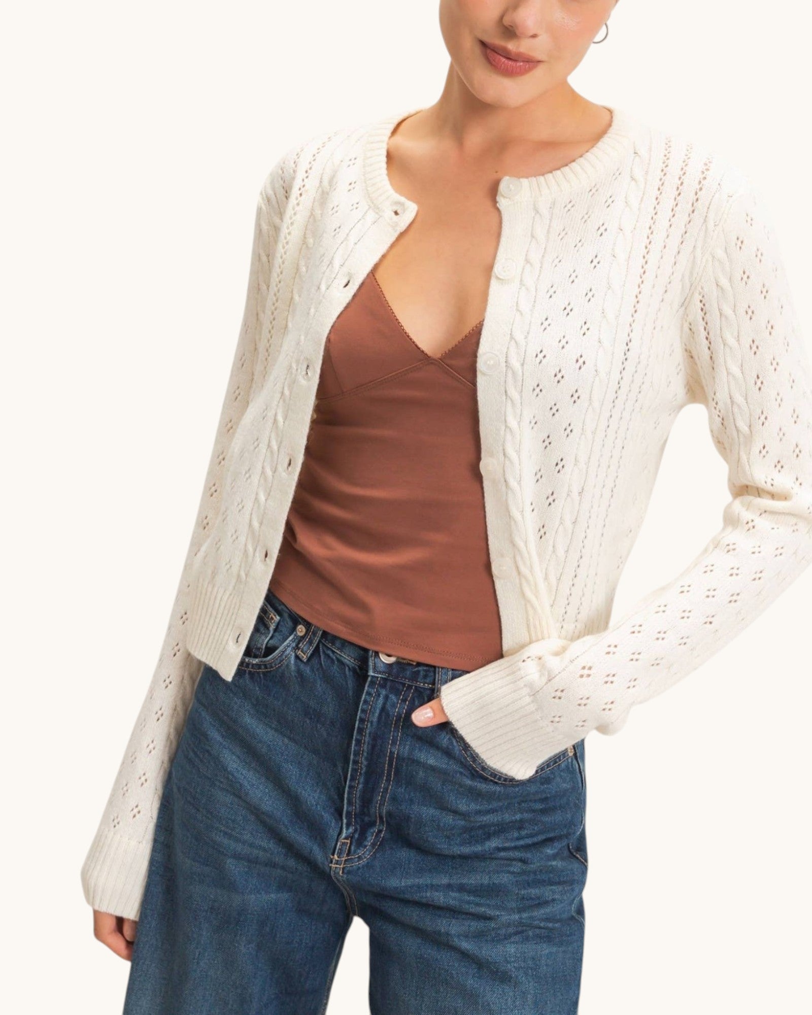 A woman wearing a cream cable-knit cardigan with diamond cutout patterns over a brown camisole top and blue jeans, shown from the chin down against a plain light background.