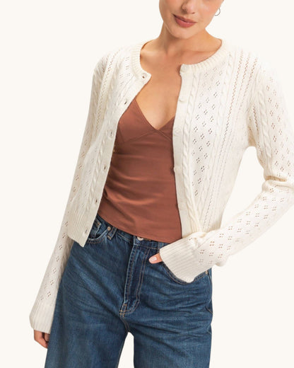 A woman wearing a cream cable-knit cardigan with diamond cutout patterns over a brown camisole top and blue jeans, shown from the chin down against a plain light background.