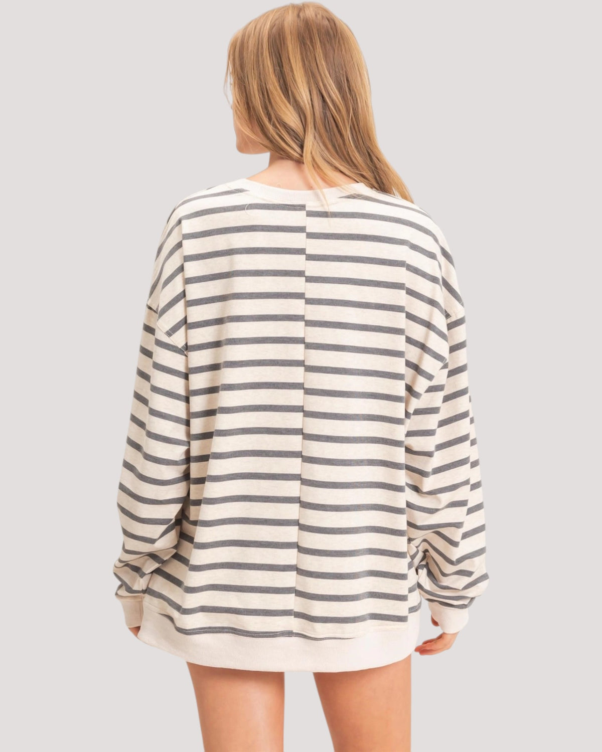 A woman shown from the back wearing an oversized long-sleeve striped top with horizontal beige and dark gray stripes, set against a solid light gray background.