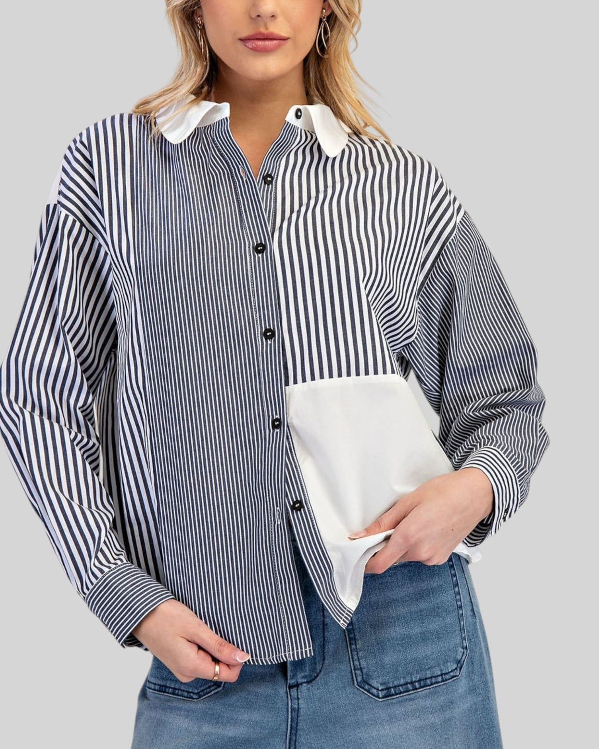 A woman wearing a long-sleeve button-up shirt with a charocal and white vertical stripe pattern, contrasting solid white collar and pocket, paired with blue jeans against a solid gray background.