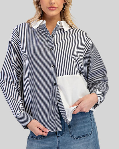 A woman wearing a long-sleeve button-up shirt with a charocal and white vertical stripe pattern, contrasting solid white collar and pocket, paired with blue jeans against a solid gray background.