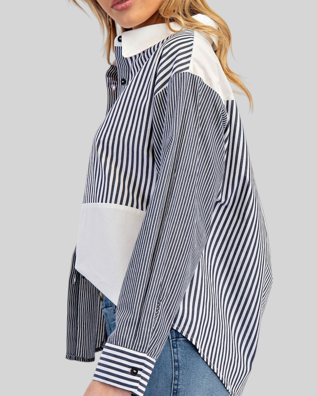 A woman wearing a long-sleeve color-block shirt with charcoal and white vertical stripes on one half, solid white on the other half, and a white collar, paired with blue jeans against a solid gray background.