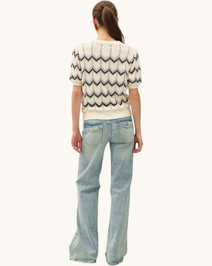 A woman shown from the back wearing a cream short-sleeve knit sweater with a multi-color zigzag pattern and light-wash blue jeans, against a plain off-white background.