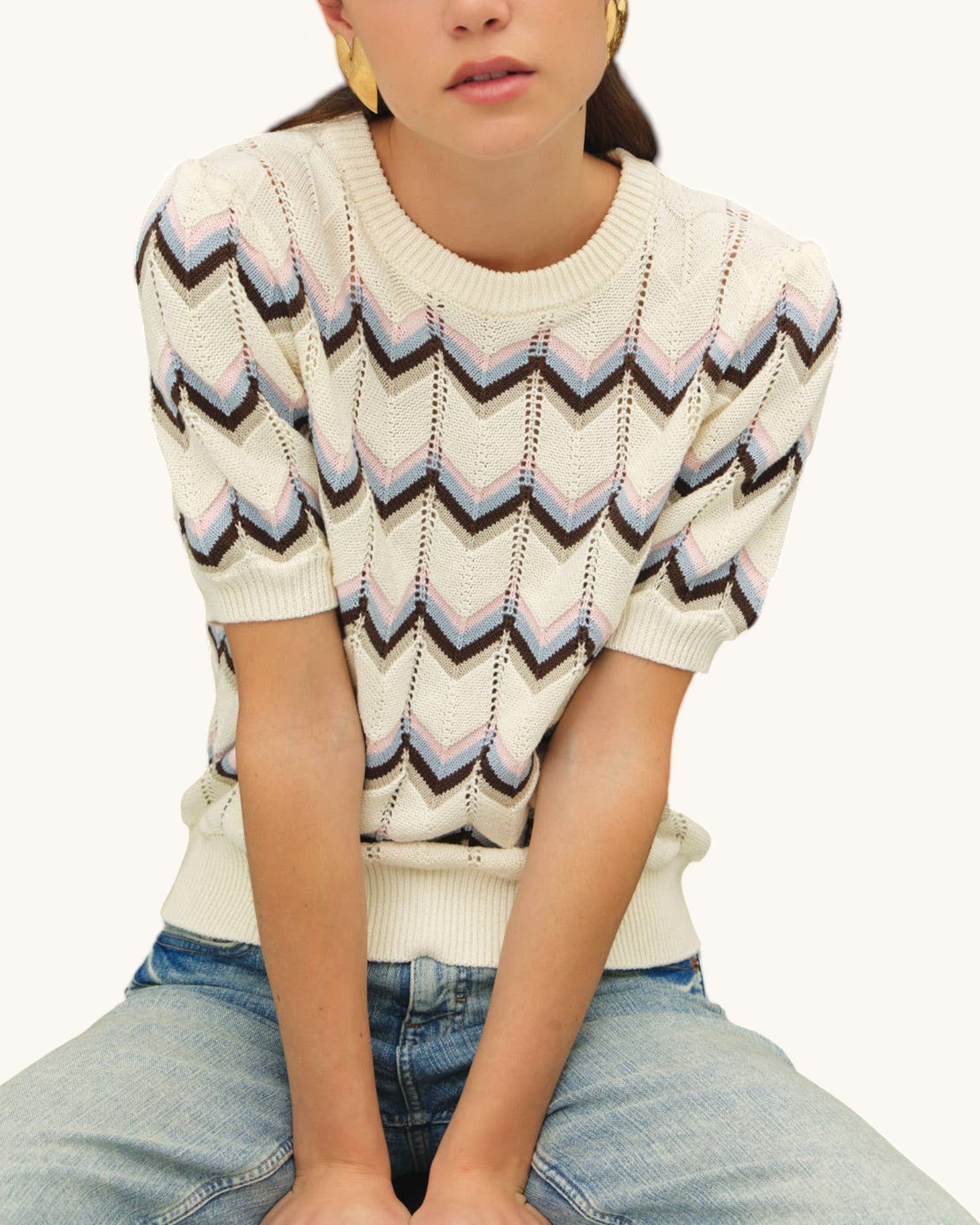 A woman wearing a cream-colored short-sleeve knit sweater with a multicolor chevron pattern in taupe, brown, light blue, and pink, paired with light-wash blue jeans.