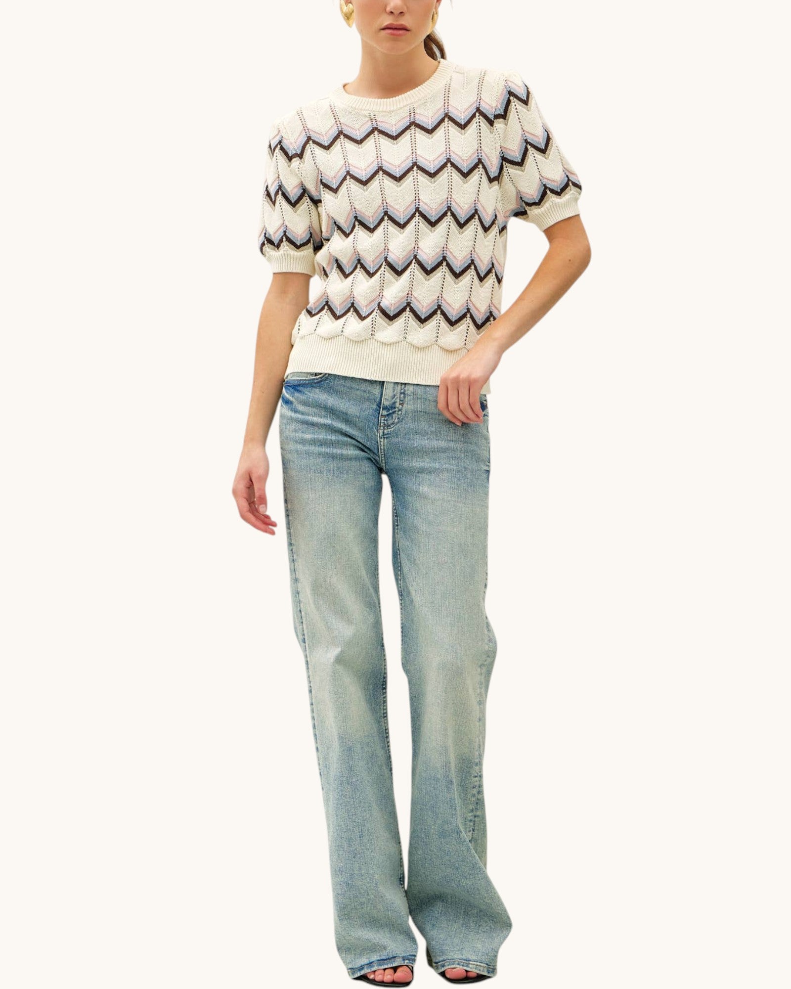 A model wearing a cream-colored short-sleeve knit sweater with a multi-color chevron pattern, paired with light-wash blue jeans, against a solid off-white background.