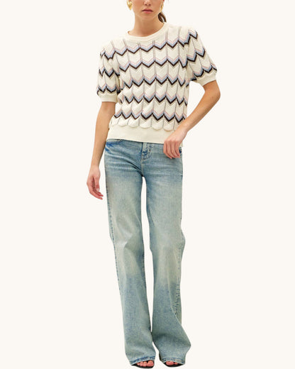 A model wearing a cream-colored short-sleeve knit sweater with a multi-color chevron pattern, paired with light-wash blue jeans, against a solid off-white background.