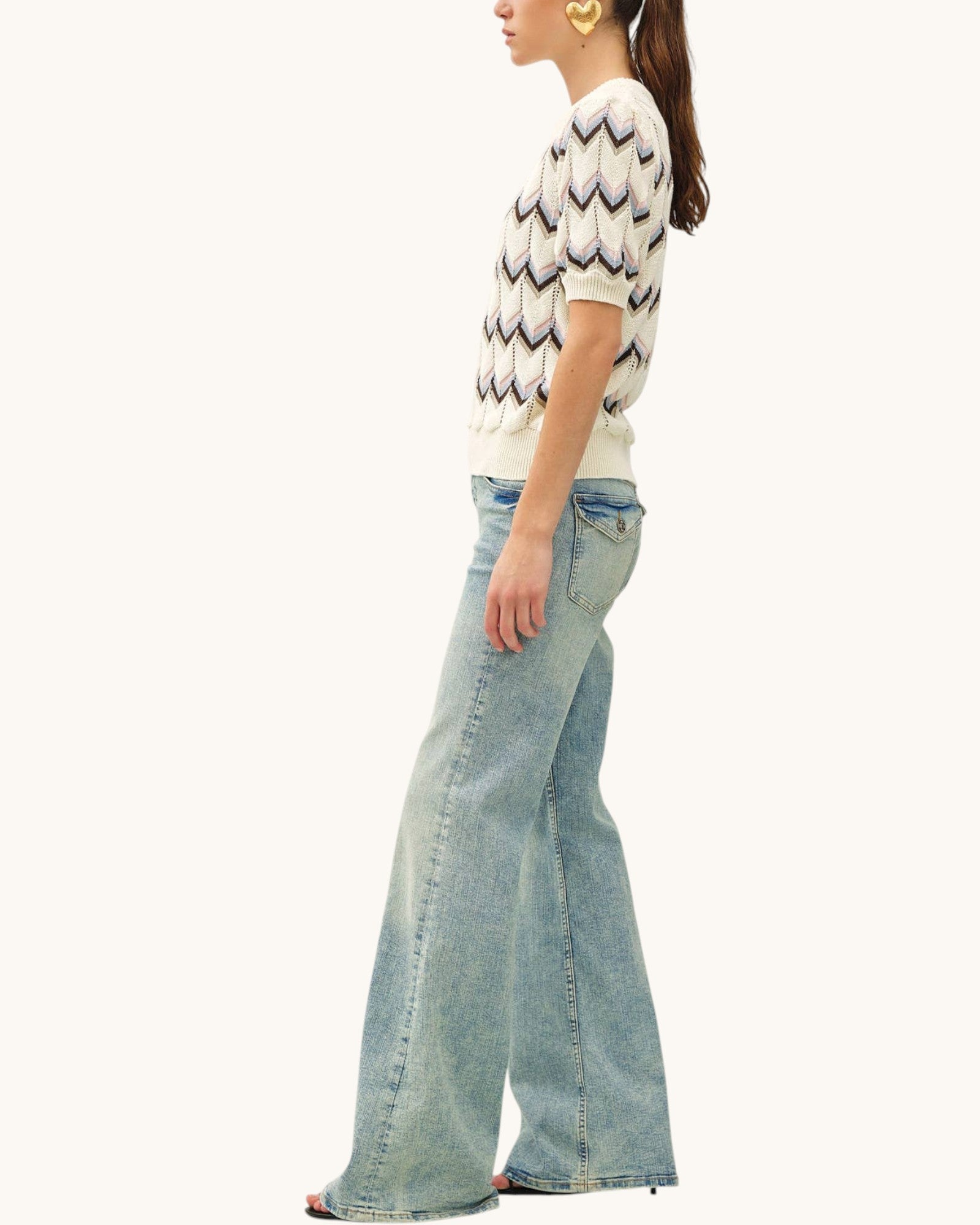 A woman shown in side profile wearing a cream knit sweater with a multi-color stripe and chevron pattern and light-wash wide-leg denim jeans, against a plain off-white studio background.