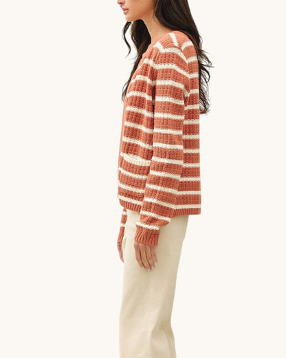A woman shown in side profile wearing a rust-orange and cream striped knit sweater cardigan with long sleeves, paired with cream flared pants and black heels, against a solid off-white background.