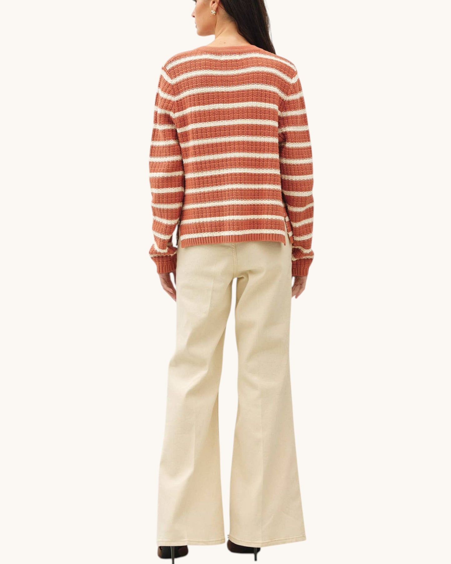 A woman shown from the back wearing a rust-orange and cream horizontally striped knit sweater and cream wide-leg trousers, against a plain off-white background.