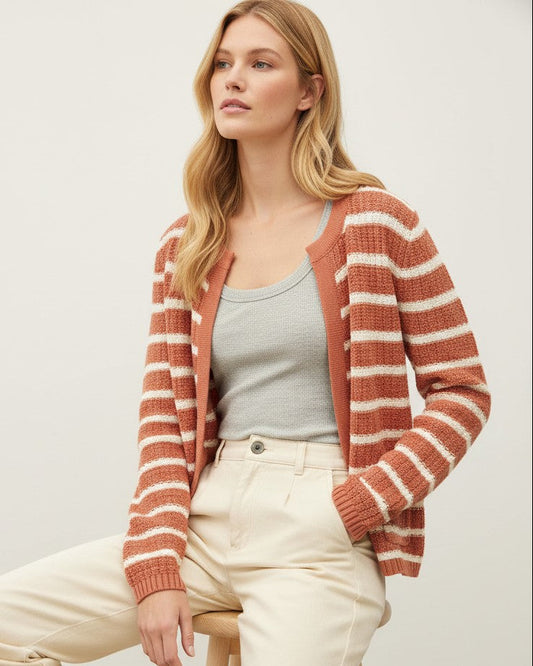 A woman seated on a wooden stool wearing a rust-orange and cream striped knit cardigan over a light gray tank top and cream pants, photographed against a plain light beige studio background.