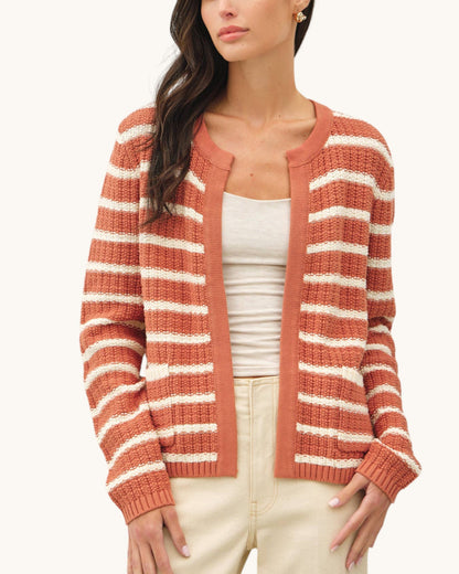 A woman wearing a rust-orange and cream striped knit cardigan over a white tank top and cream pants, shown from the head down against a plain light background.