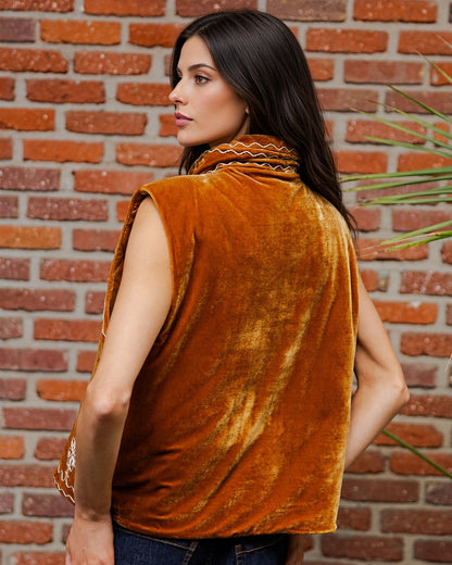 Back view of a woman wearing a sleeveless velvet vest in a rich burnt orange color, standing in front of a red brick wall.