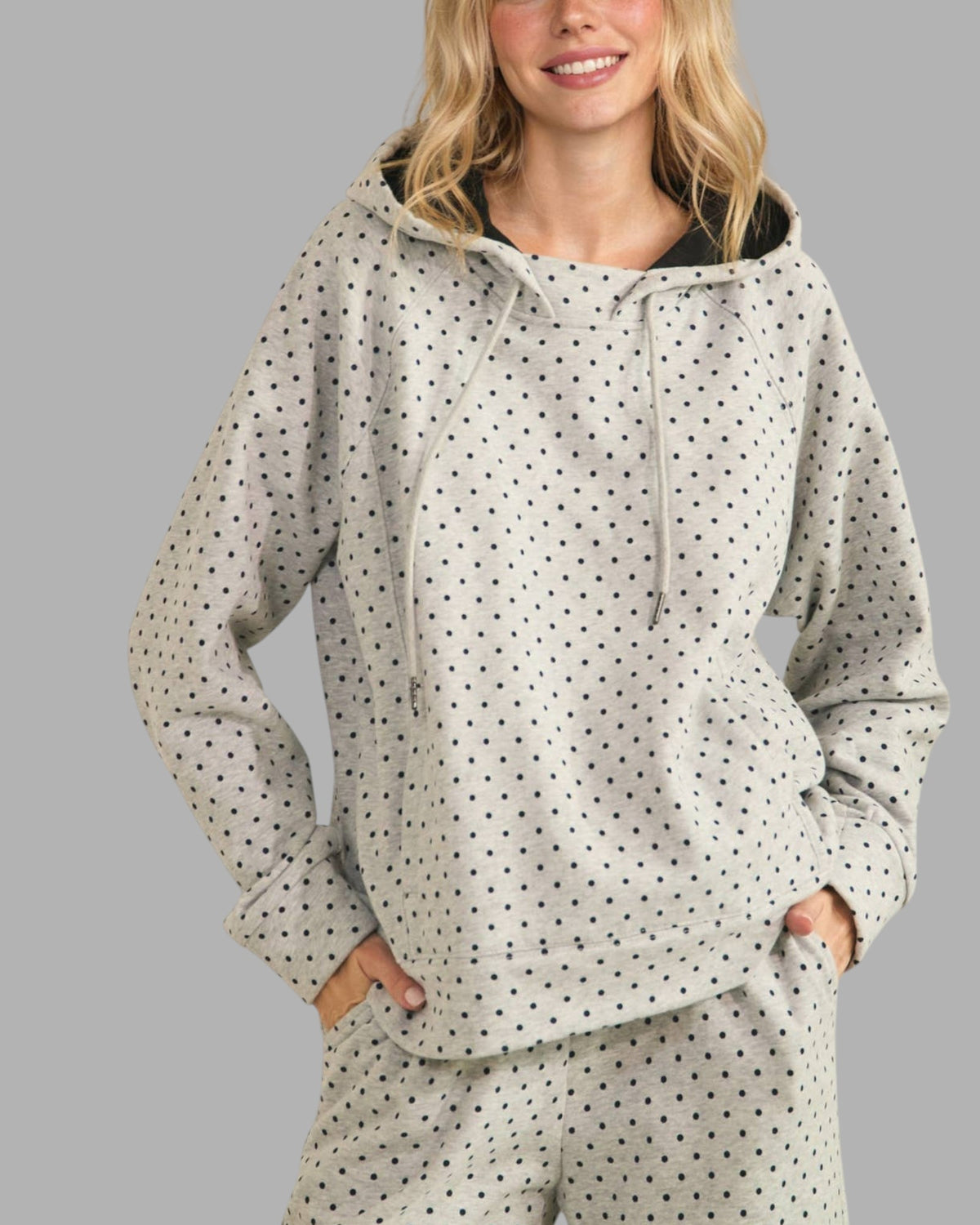 A woman wearing a light gray hooded sweatshirt and matching sweatpants with a small black polka dot pattern, posed against a solid gray background.