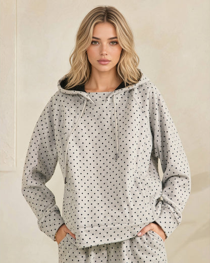 A woman modeling a light gray hooded sweatshirt with a black polka dot pattern, featuring a black-lined hood and drawstrings, set against a neutral beige background.