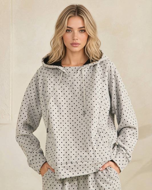 A woman modeling a light gray hooded sweatshirt with a black polka dot pattern, featuring a black-lined hood and drawstrings, set against a neutral beige background.