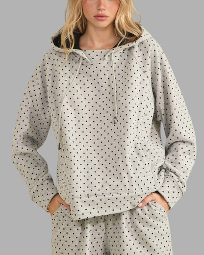 A woman wearing a light gray hooded sweatshirt with a black polka dot pattern, paired with matching jogger pants, against a solid gray background.