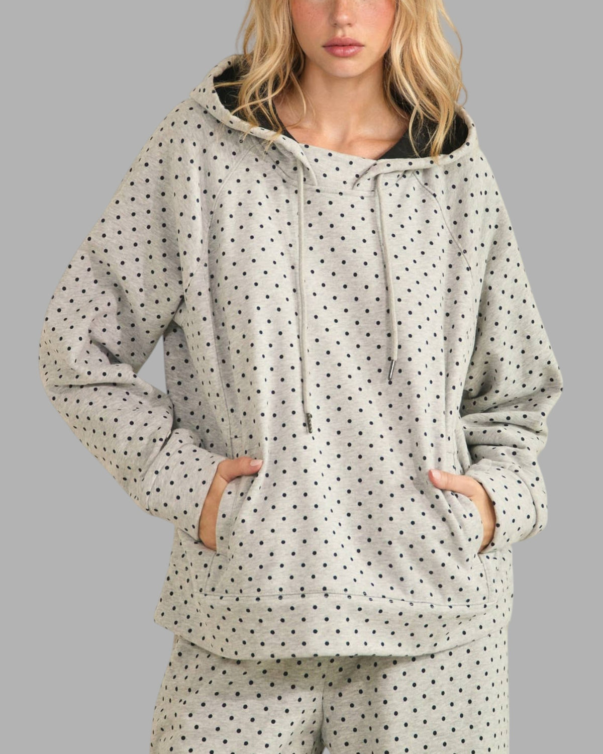A woman wearing a light gray hooded sweatshirt and matching pants set, both covered in a small black polka dot pattern. The hoodie features a black-lined hood with white drawstrings.