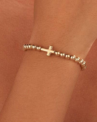 A delicate gold beaded bracelet with a central cross charm, shown worn on a person's wrist against a warm skin-toned background.