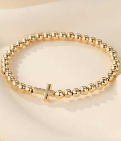 A gold beaded bracelet with a small cross charm, displayed on a soft cream-colored surface.