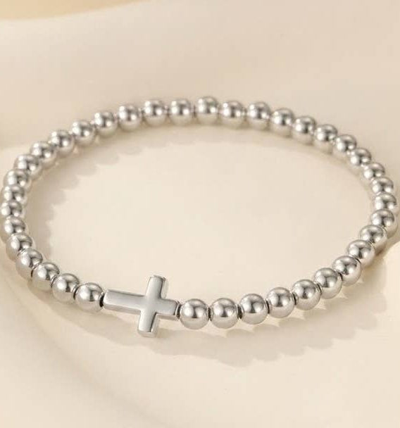 A silver beaded stretch bracelet with a polished cross charm, displayed on a soft beige background.
