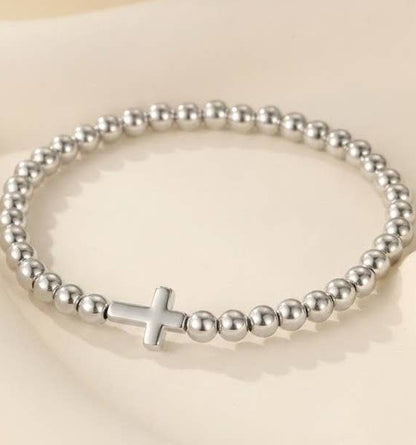 A silver beaded stretch bracelet with a polished cross charm, displayed on a soft beige background.